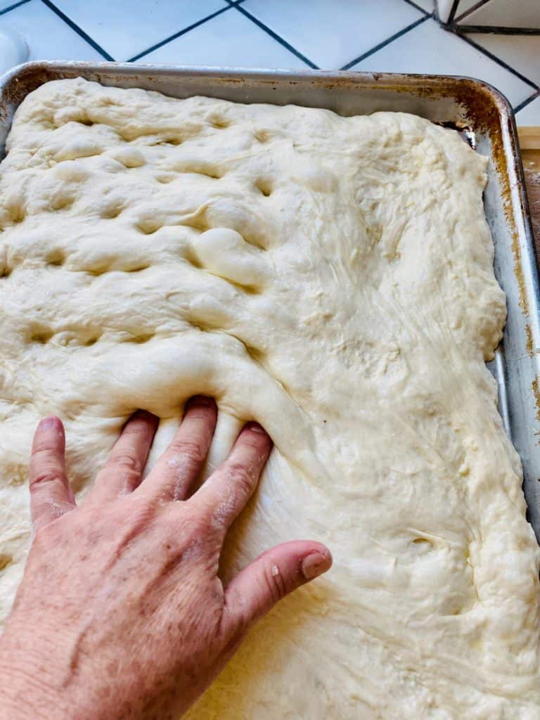 Fingers of a left hand making dimples in focaccia dough