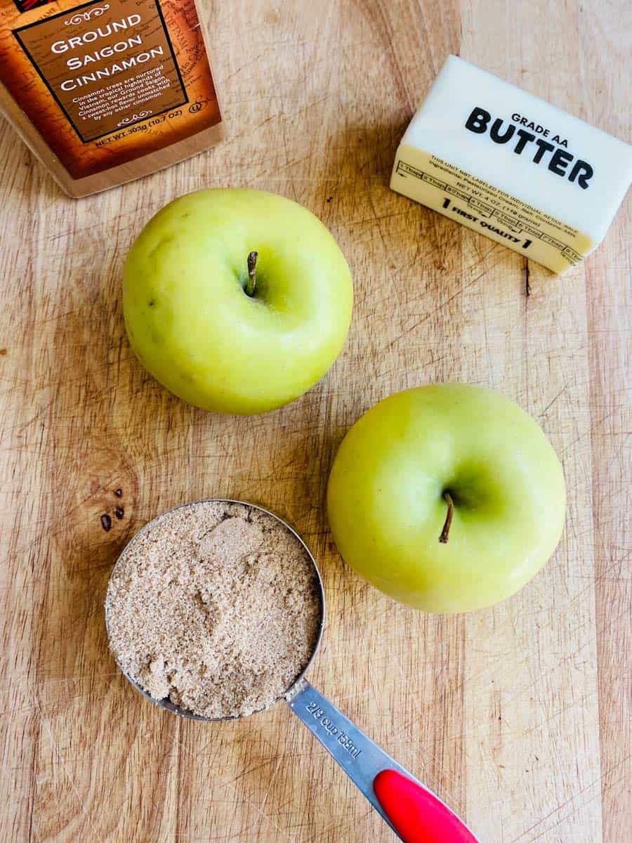 Two Golden Delicious apples, cup of brown sugar, stick of butter, and jar of cinnamon on a wooden cutting board.