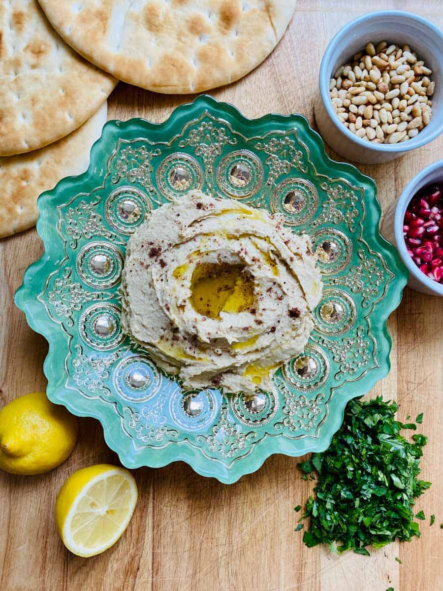 Plate of hummus, pita, cut lemons, and piles of parsley, pine nuts, and pomegranate seeds.