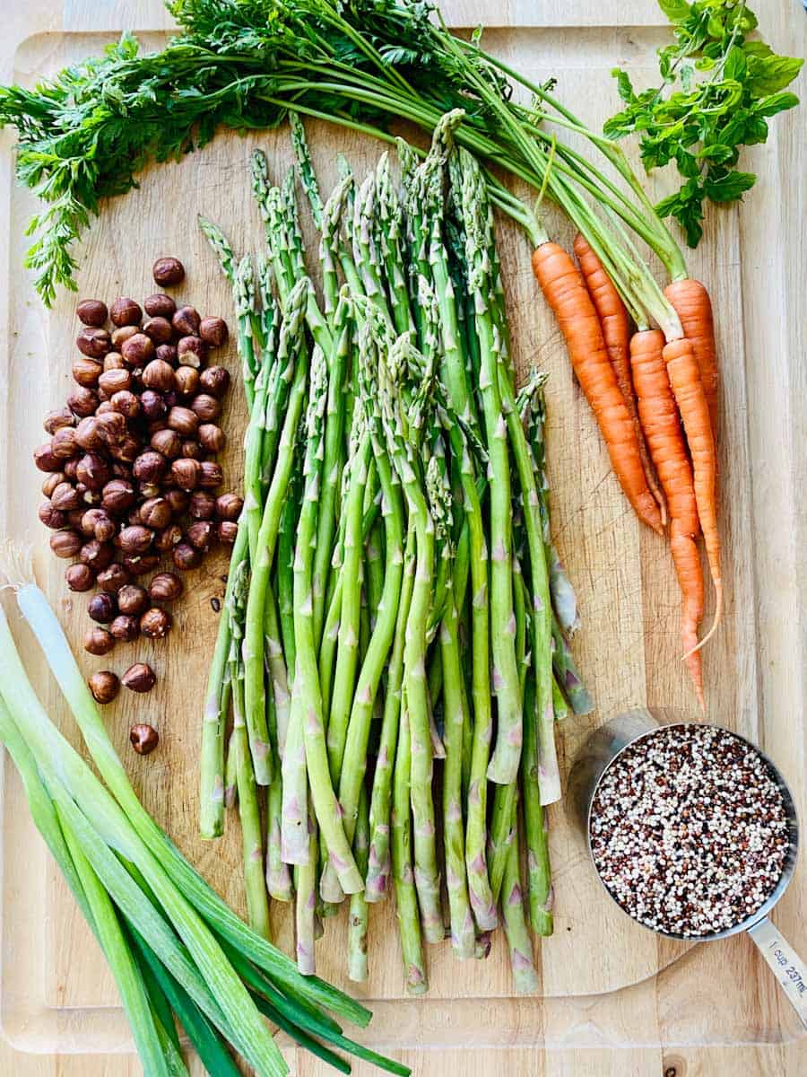 Quinoa, asparagus, green onions, hazelnuts, herbs, and carrots laid out on a wooden board.