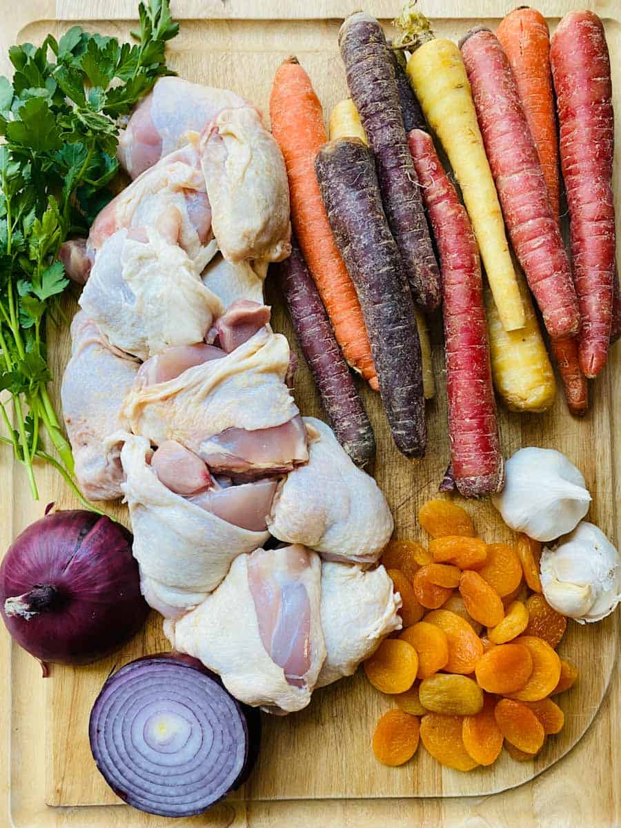 Chicken, carrots, garlic, dried apricots, red onion, and parsley on a cutting board.