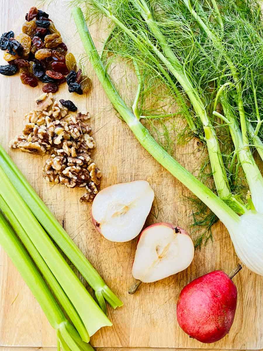 Fennel, red pears, celery, walnuts, and raisins on a wooden cutting board.