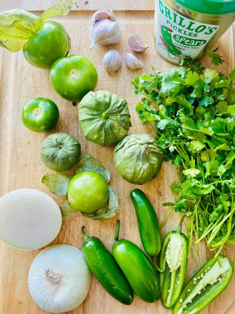 Tomatillos, garlic, pickles, cilantro, jalapeno, and onion on a cutting board.