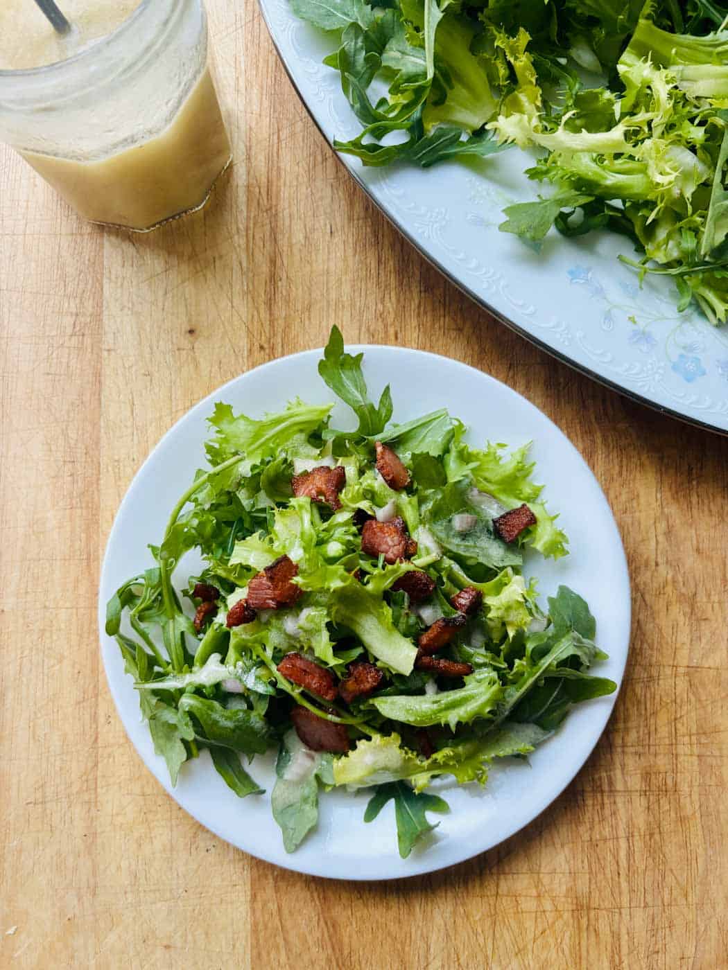 A jar of dressing, plate of greens, and composed salad on a cutting board.